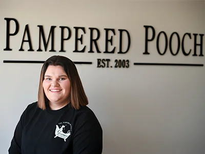 Lydia Sullivan, groomer at The Pampered Pooch, smiling in front of the business name and established date wall sign.