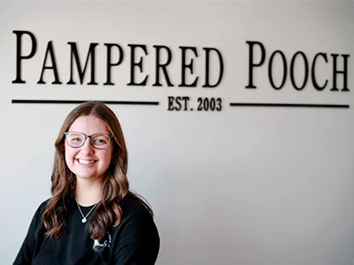 Lexie Vogel, a groomer at The Pampered Pooch, smiling in front of the business name and established date wall sign.