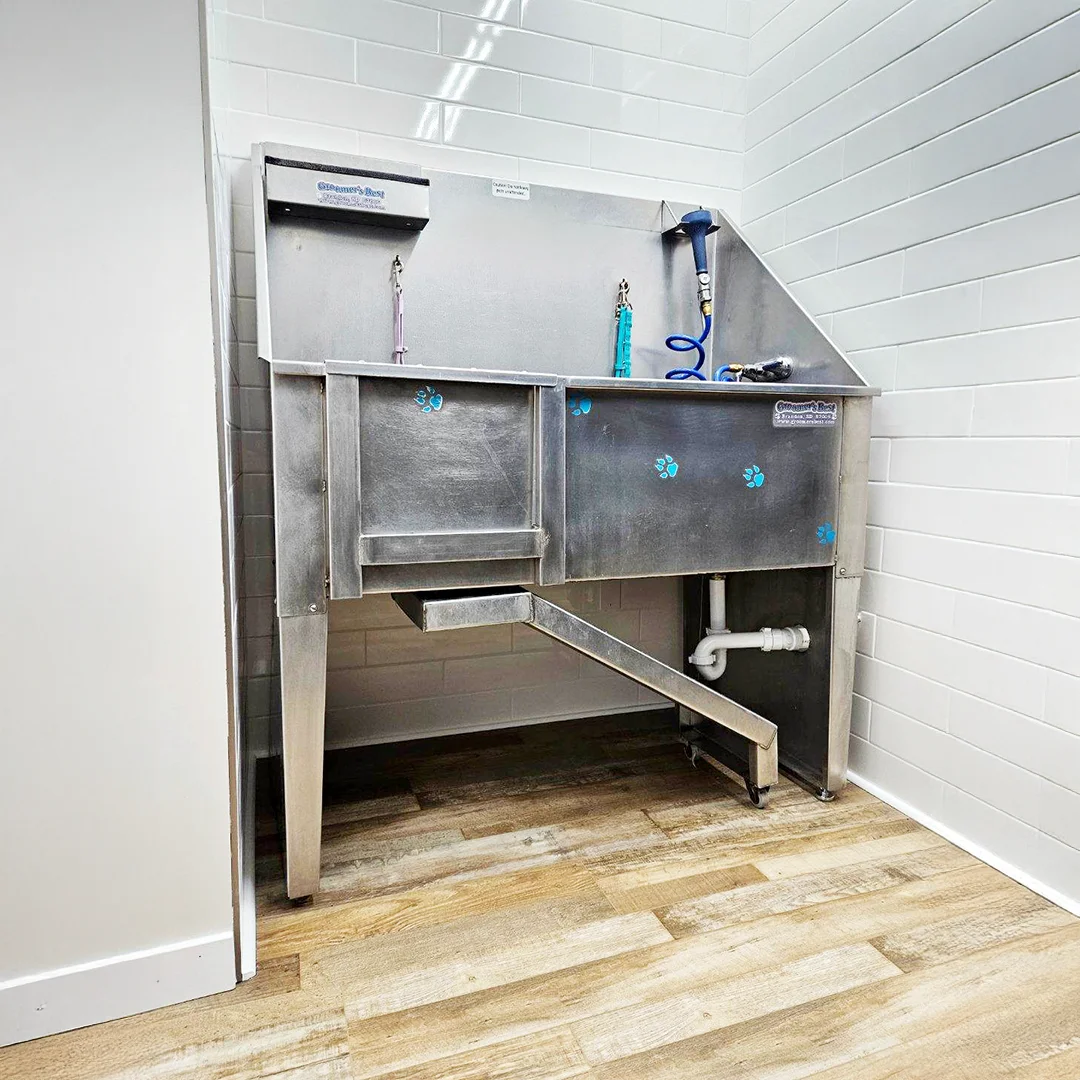 Professional stainless steel grooming tub with hose attachments, safety hooks, and a ramp for easy pet access inside The Pampered Pooch grooming facility.