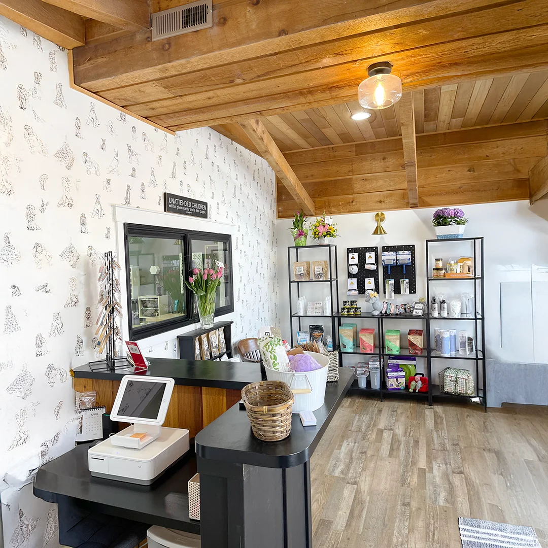 Reception area at The Pampered Pooch with a checkout counter, wooden ceiling beams, floral accents, and shelves stocked with grooming products and pet supplies.