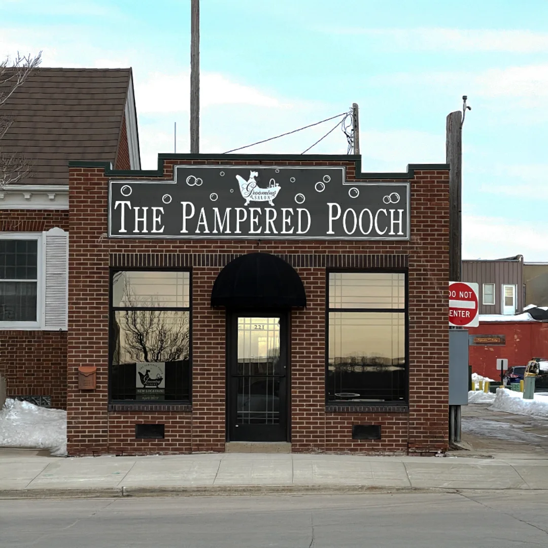 Exterior of The Pampered Pooch brick building with a black awning, large front windows, and signage displaying the business name and grooming salon logo.